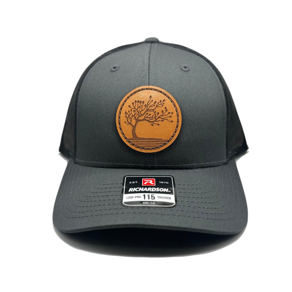 Front view of the Blossom Leather Patch Hat by Outer Wings in charcoal/black, featuring a low-profile Richardson 115 hat with a distinctive leather patch of the Blossom design. The charcoal front paired with a black mesh back provides a bold, contrasting look.