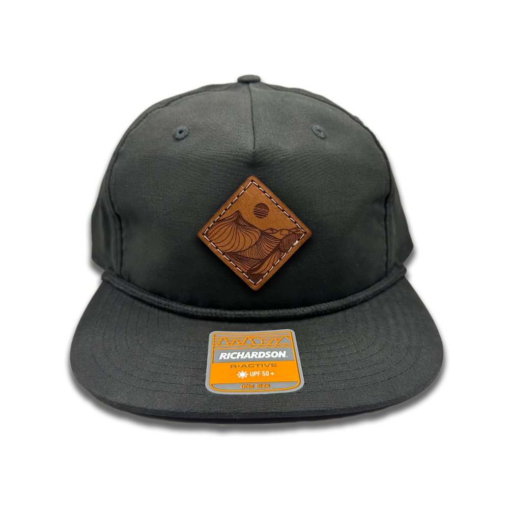 Front view of the Desert Mountain Leather Patch Hat by Outer Wings in black/black on the Richardson 256 Umpqua Rope Hat. This hat showcases a circular leather patch with the Desert Mountain design, laser engraved and hand-sewn for durability, complemented by a flat bill, rope detail, and an adjustable SnapBack closure.