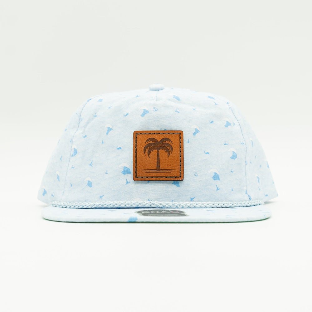 A light blue cotton flat-brim cap with a subtle blue speckled pattern, featuring a brown faux-leather patch on the front with an engraved palm tree logo. The cap is photographed against a clean, off-white studio background.

Color: Blue Wave
