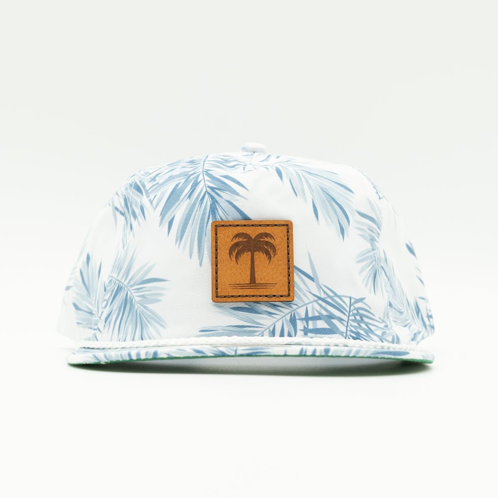 A white snapback cap with a light blue palm leaf pattern and a brown faux-leather patch on the front featuring a palm tree logo.

Color: Floral Mist