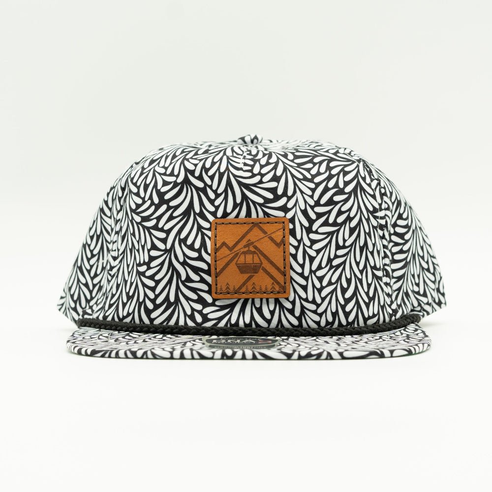 A flat-brim baseball cap with a black and white leaf pattern, featuring a brown leather patch on the front with a cable car graphic.

Color: Throwback Black