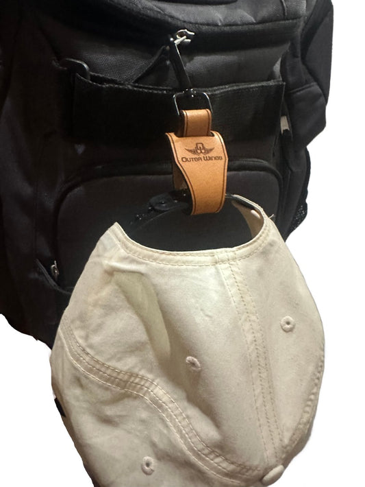 Leather hat clip by Outer Wings attached to a backpack with a hat securely hanging from it. The clip features a strong leather loop and a 360-degree swivel quick link keychain, demonstrating its durability and functionality for convenient hat carrying.