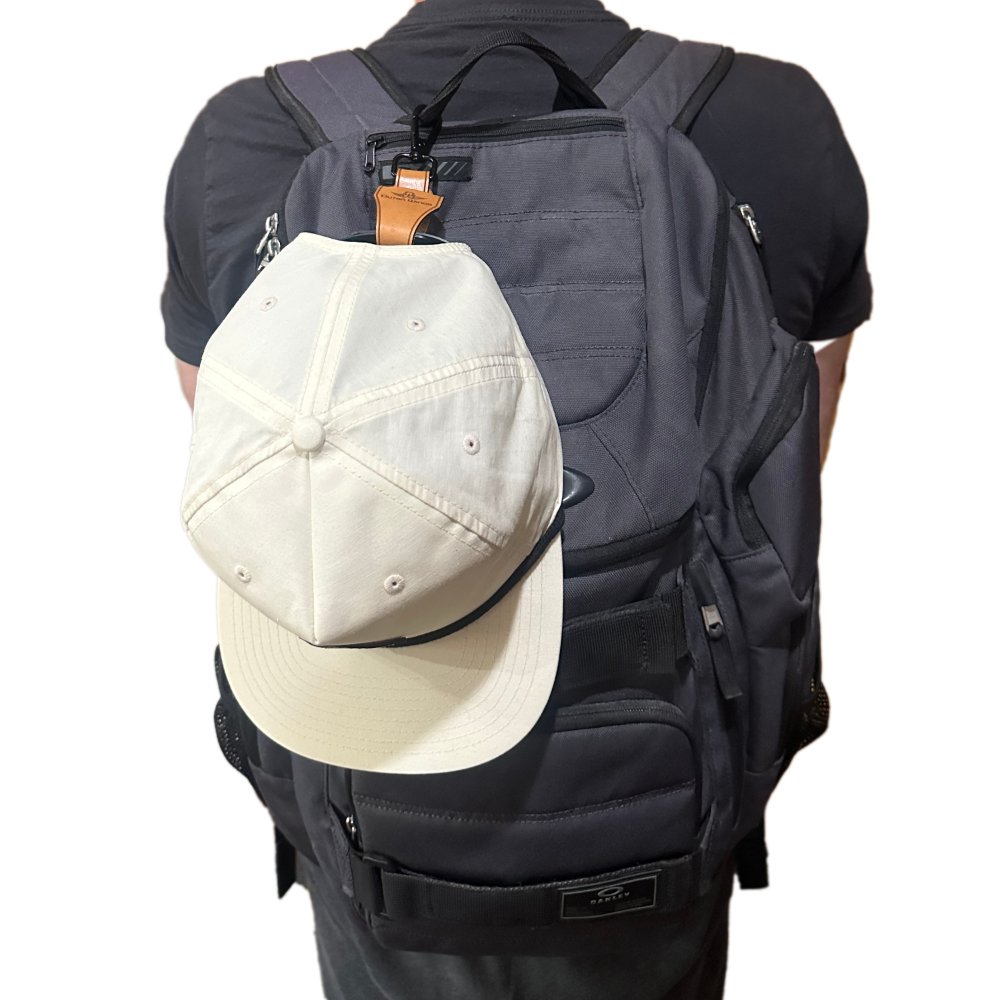 Close-up of the Outer Wings leather hat clip with a hat hanging from a backpack, highlighting the thick leather loop and 360-degree swivel quick link keychain. This stylish accessory offers a secure and practical way to carry hats on the go