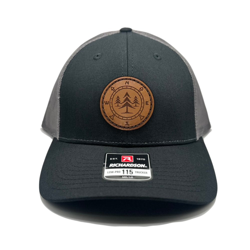 Front view of the Lost Pine Hat in Black/Charcoal by Outer Wings, showcasing a circle-shaped leather patch with a compass and pine trees design. Built on the Richardson 115 Low Profile Hat, it combines style and comfort with its structured six-panel design and adjustable snapback.