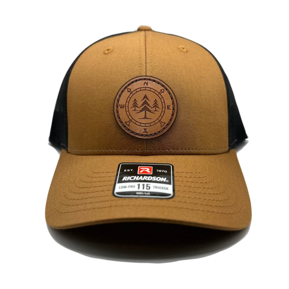 Front view of the Lost Pine Hat in Caramel/Black by Outer Wings, featuring a circle-shaped leather patch with a compass and pine trees design. Built on the Richardson 115 Low Profile Hat, it offers a structured six-panel design with a comfortable and adjustable snapback closure.