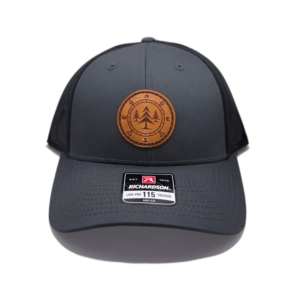 Front view of the Lost Pine Hat in Charcoal/Black by Outer Wings, showcasing a circle-shaped leather patch with a compass and pine trees design. The Richardson 115 Low Profile Hat combines style and function with a structured six-panel build and adjustable snapback closure.