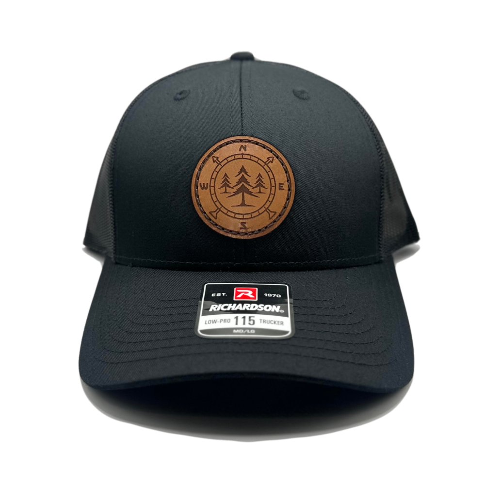 Front view of the Lost Pine Hat in Black/Black by Outer Wings, featuring a circle-shaped leather patch with a compass and pine trees design. This hat is crafted on the Richardson 115 Low Profile Hat with a cotton/polyester blend, 100% polyester mesh back, and adjustable snapback closure.