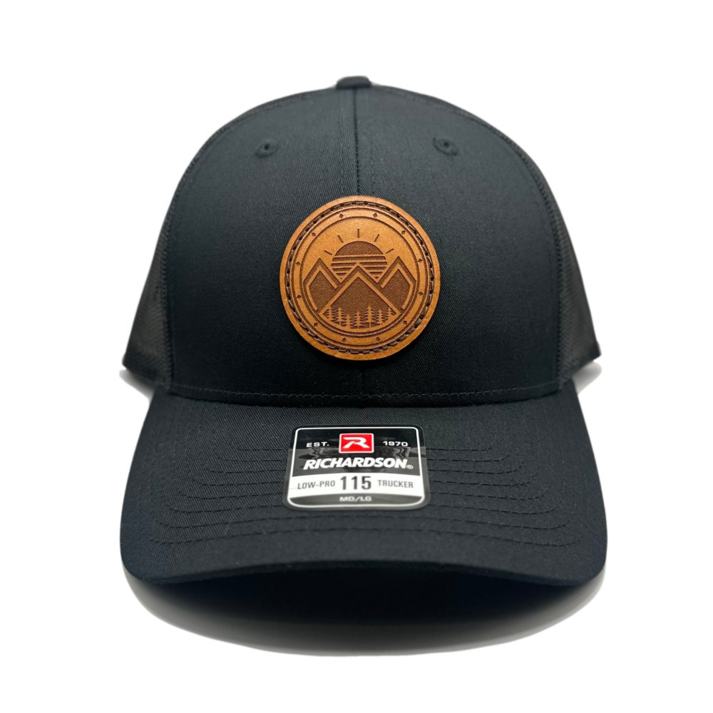 Front view of the Modern Mountains Leather Patch Hat in Black/Black by Outer Wings, crafted on the Richardson 115 Low Profile Hat. It features a genuine leather patch with a laser-engraved design of modern mountains and a sunset. The hat has a structured six-panel design, a pre-curved visor, and an adjustable SnapBack closure.