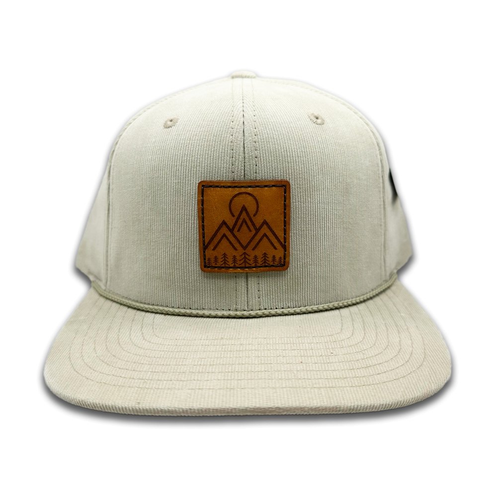 Front view of the Moonlite Mountain Hat in tan by Outer Wings, displaying a square leather patch with a laser-engraved design of mountains, pine trees, and a moon. Built on the Richardson 253 with 100% cotton corduroy, this hat features a high-profile, six-panel structure and a flat bill for a modern look