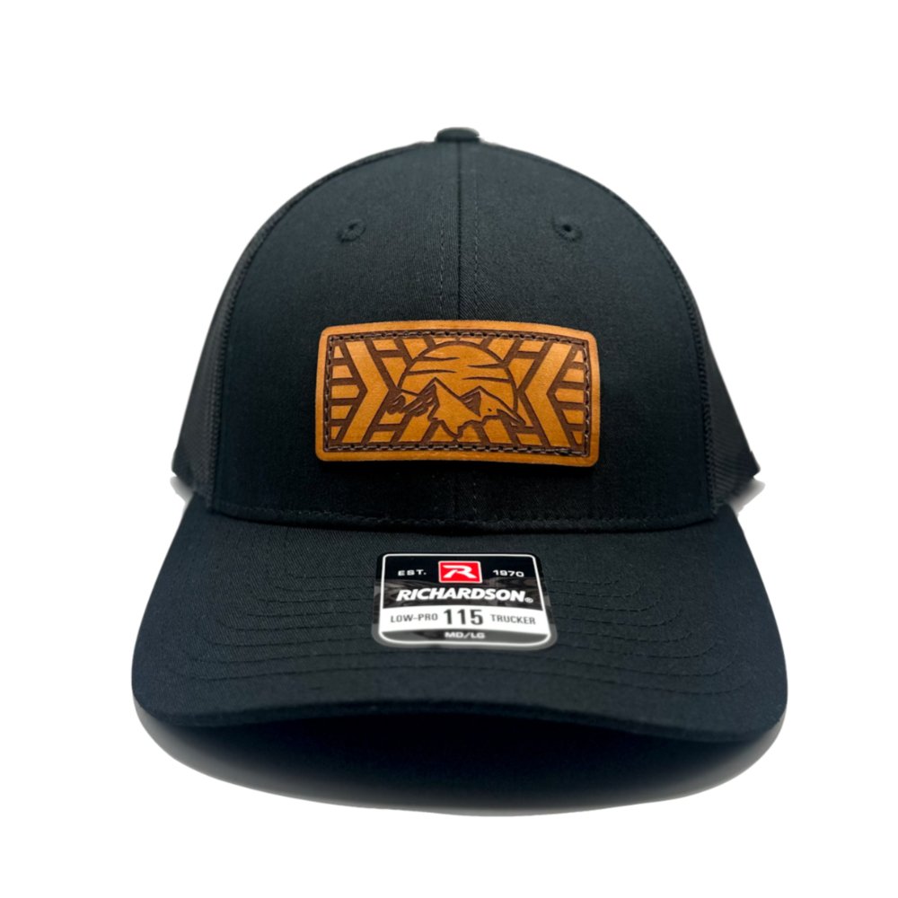 Front view of the Mosaic Mountain Leather Patch Hat by Outer Wings in black/black, crafted on a low profile Richardson 115 hat. This custom leather patch hat features a square leather patch with a laser-engraved mountain and sunset design, offering a sleek and stylish outdoors hat with a comfortable fit and adjustable SnapBack closure.