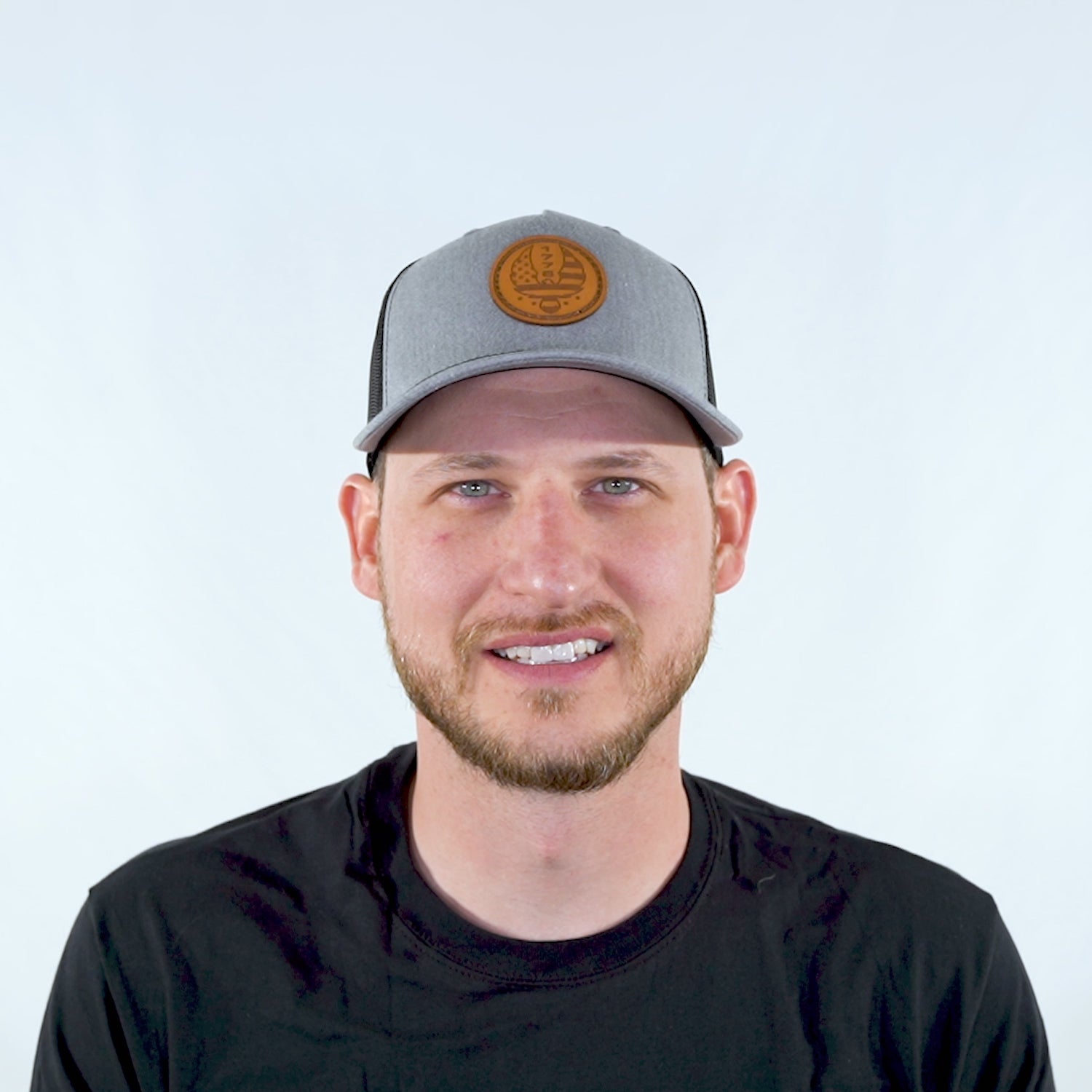 Man wearing Heather Grey/Black Richardson 112FP five panel trucker hat with sewn 1776 eagle leather patch, turning his head left to right to show the patriotic leather patch hat fit and mesh back.