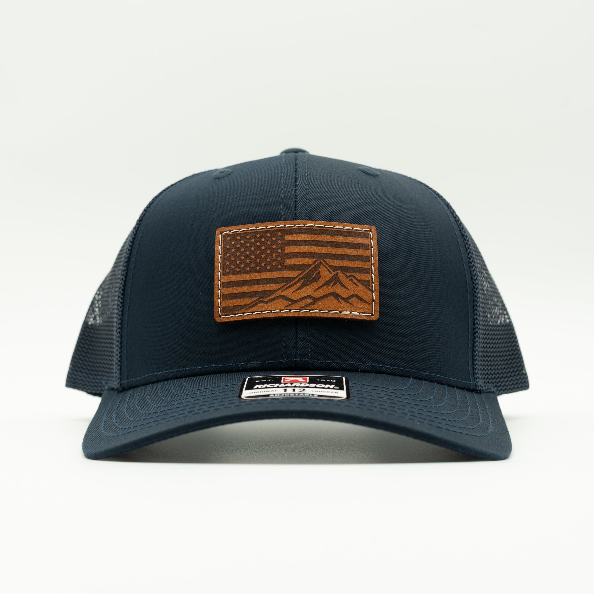 A dark navy blue trucker hat with a brown leather patch on the front featuring an American flag and mountain design, photographed against a clean white background.

Color: Navy/Navy