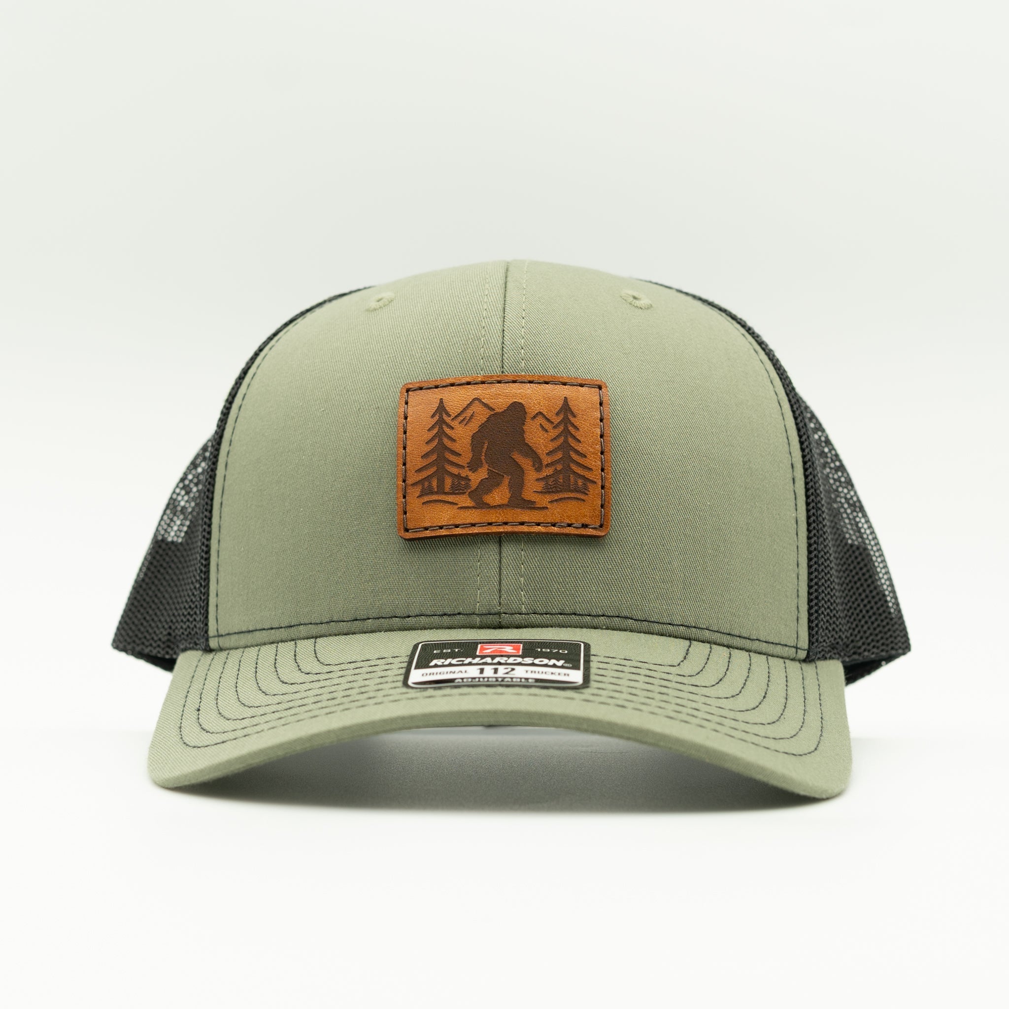 Front view of loden/black Mountain Myth Bigfoot leather patch trucker hat, Richardson 112 mid-profile fit for everyday outdoor wear.