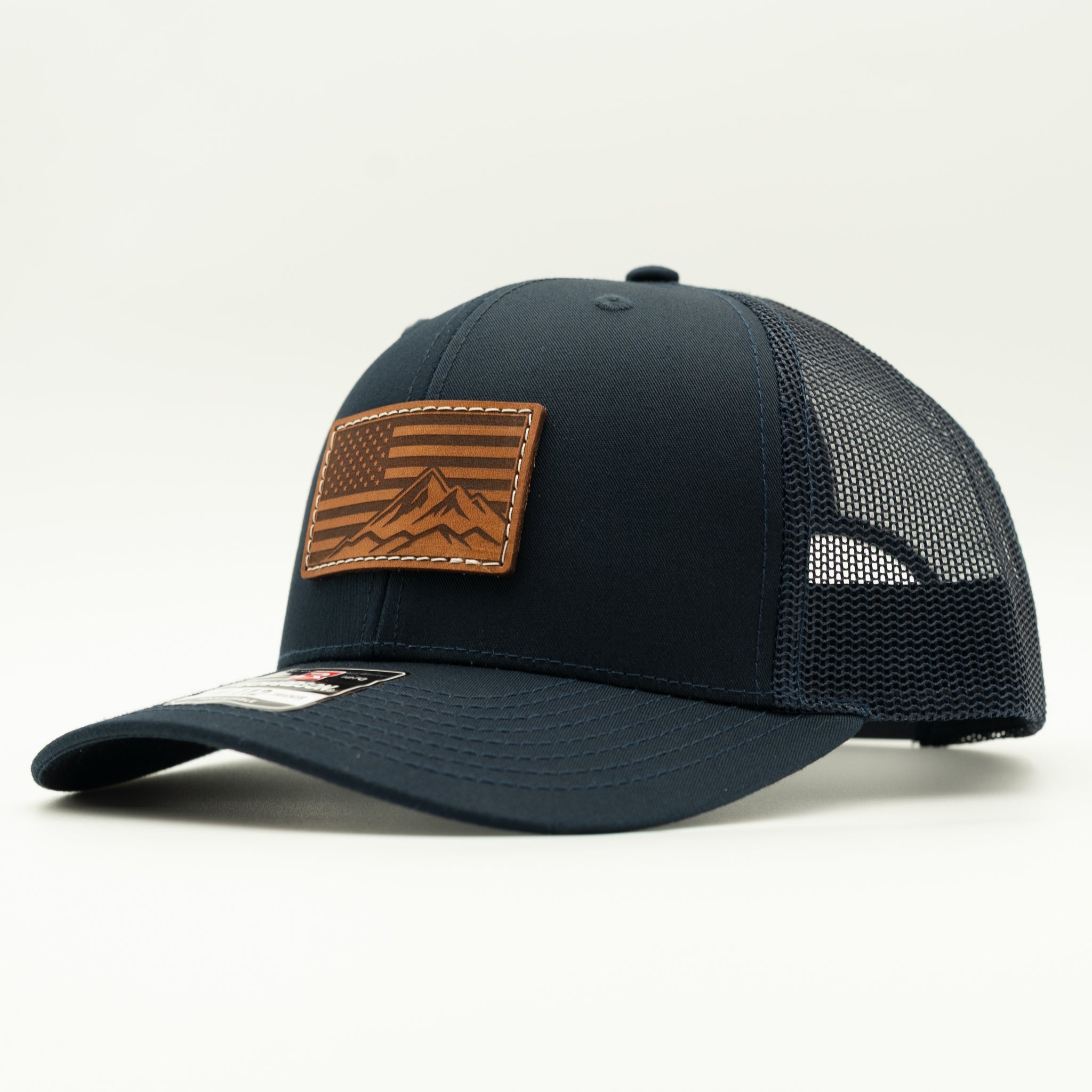 A dark navy blue trucker hat with a brown leather patch on the front featuring a mountain and American flag design, set against a plain white background.

Color: Navy/Navy