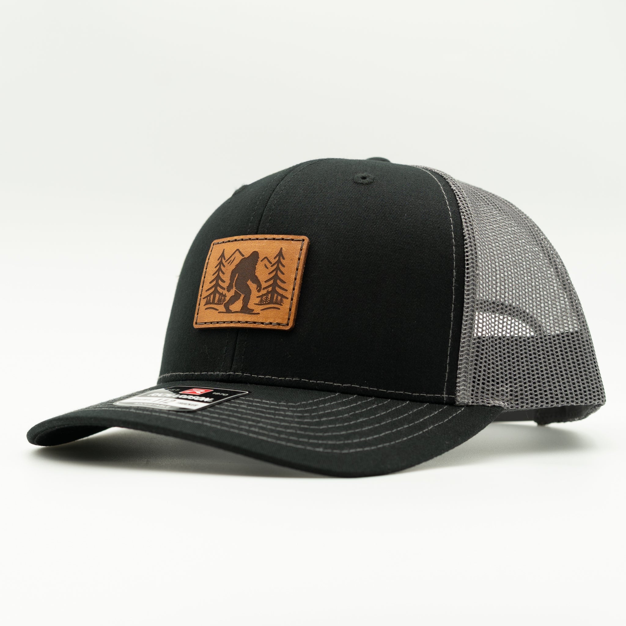Side profile of black front, charcoal mesh Richardson 112 Mountain Myth hat featuring engraved Bigfoot leather patch and curved bill.