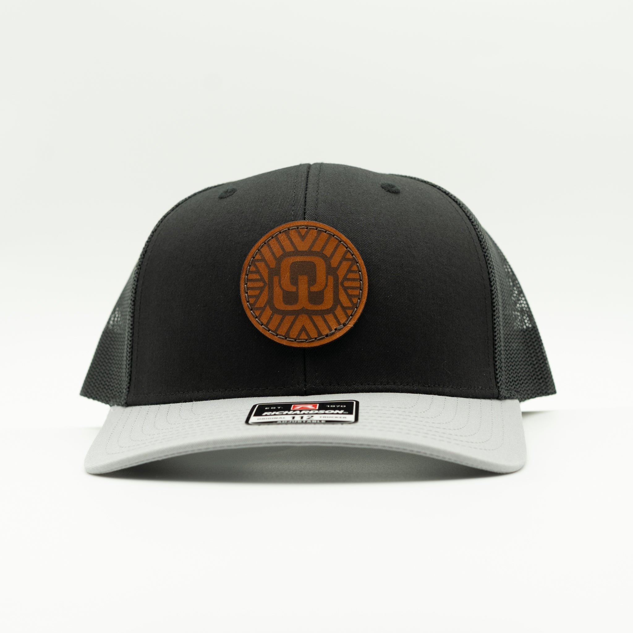 A black and gray trucker cap with a brown circular leather patch on the front, photographed against a clean white background.

Color: Black/Grey Combo