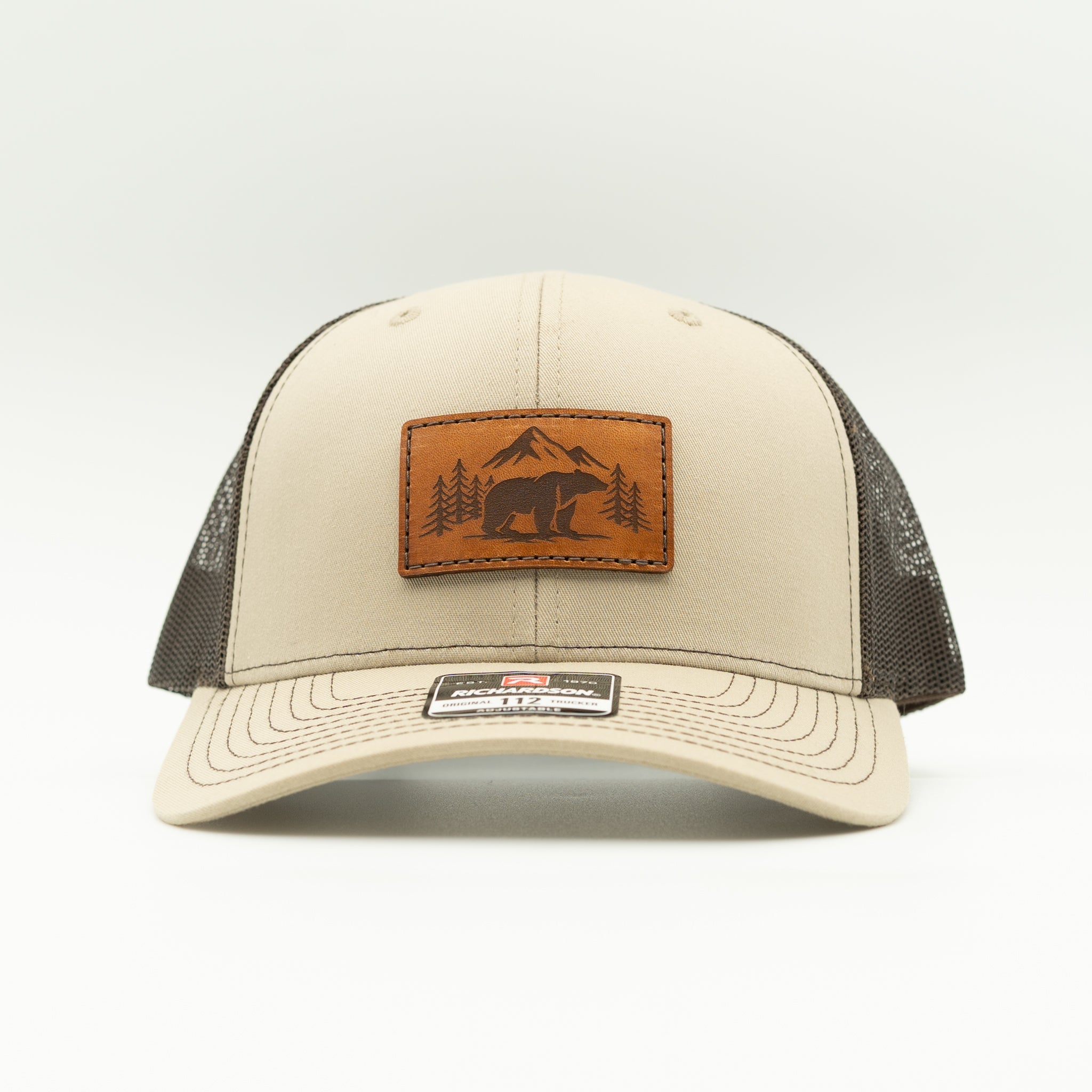 A beige trucker-style cap with dark mesh back panels and a brown leather patch on the front featuring a bear and mountain design. A small rectangular brand tag is visible on the brim.

Color: Khaki/Coffee