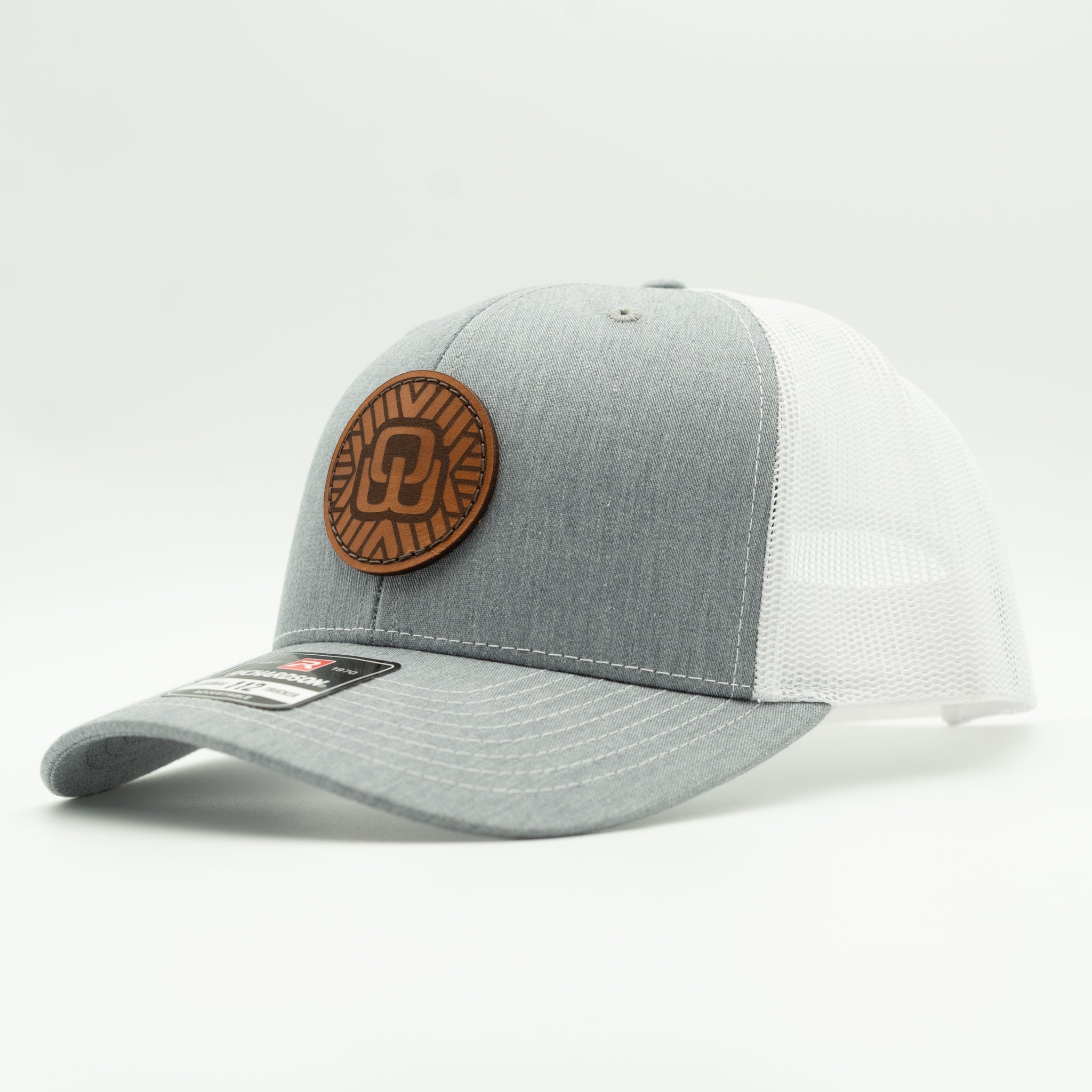 A gray and white trucker cap with a brown circular leather patch on the front, photographed on a clean white background.

Color: Heather Grey/White