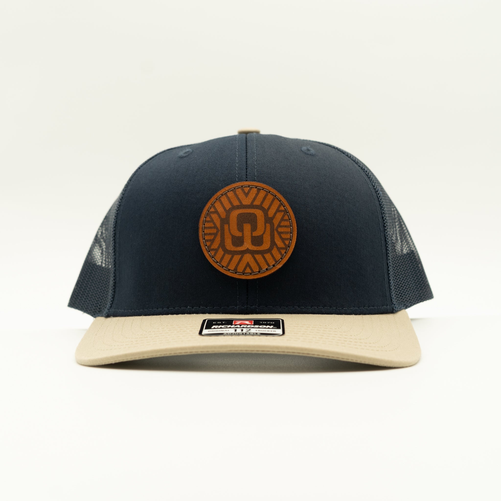 A navy blue trucker hat with a beige flat brim and a brown circular leather patch on the front. The patch features a moasic and OW for outer wings, and a small black and white brand label is visible on the brim.

Color: Navy/Khaki Combo