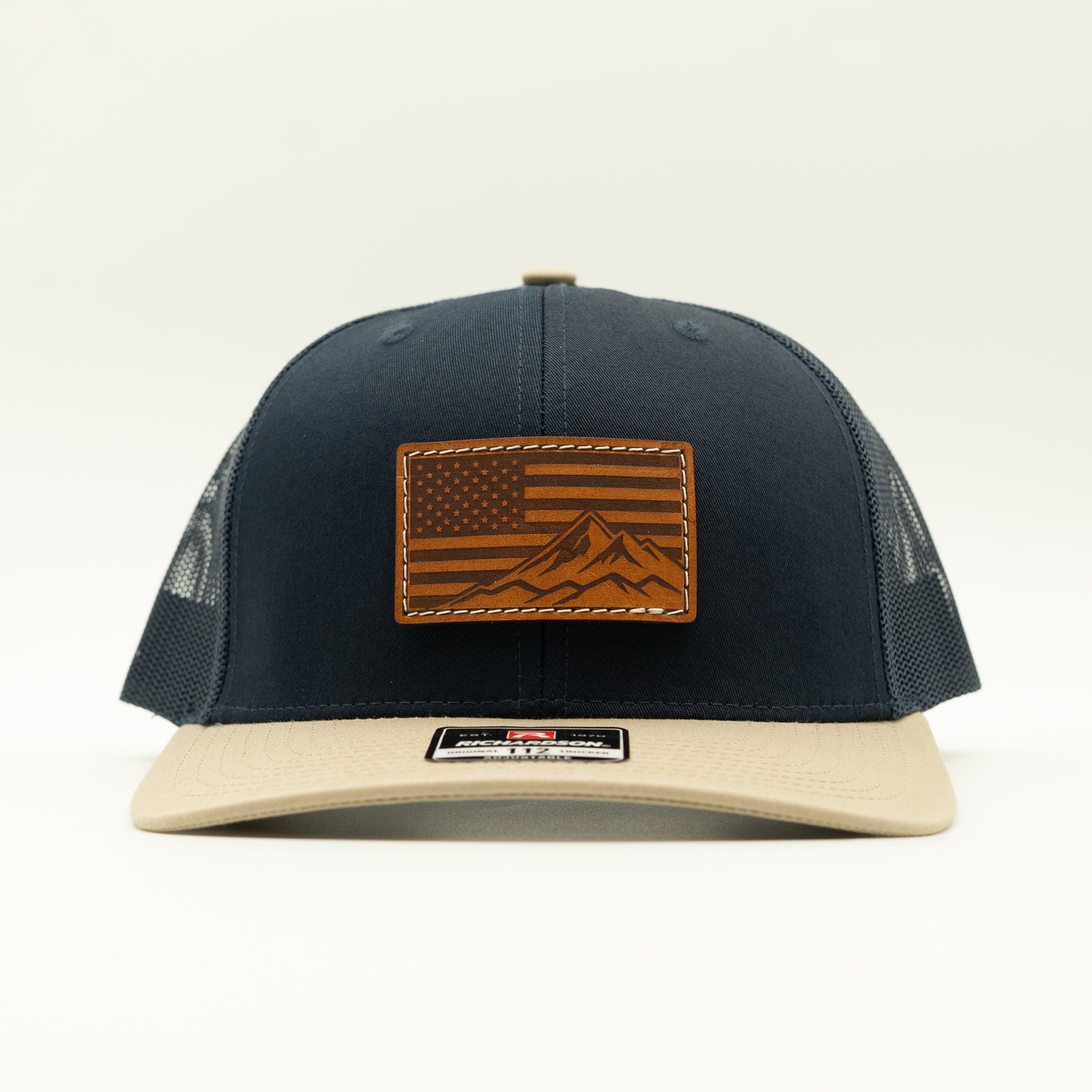 A navy blue trucker cap with a beige flat brim, featuring a brown leather patch on the front with an American flag and mountain design.

Color: Navy/Khaki Combo