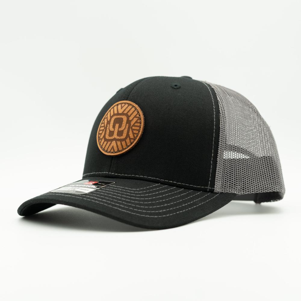 A black trucker-style baseball cap with a curved brim, black front panel, and gray mesh back panels. It features a circular brown leather patch on the front, and white contrast stitching on the brim.

Color: Black/Charcoal