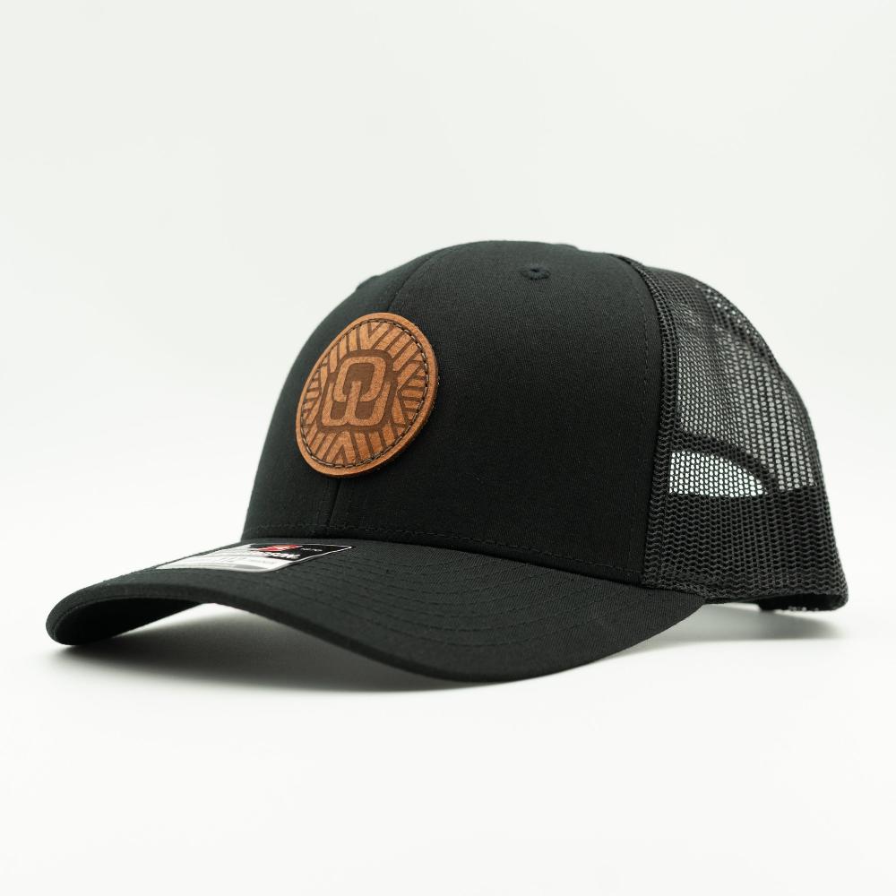 A black trucker-style baseball cap with a curved brim, black mesh back panels, and a circular brown leather patch on the front.

Color: Solid Black