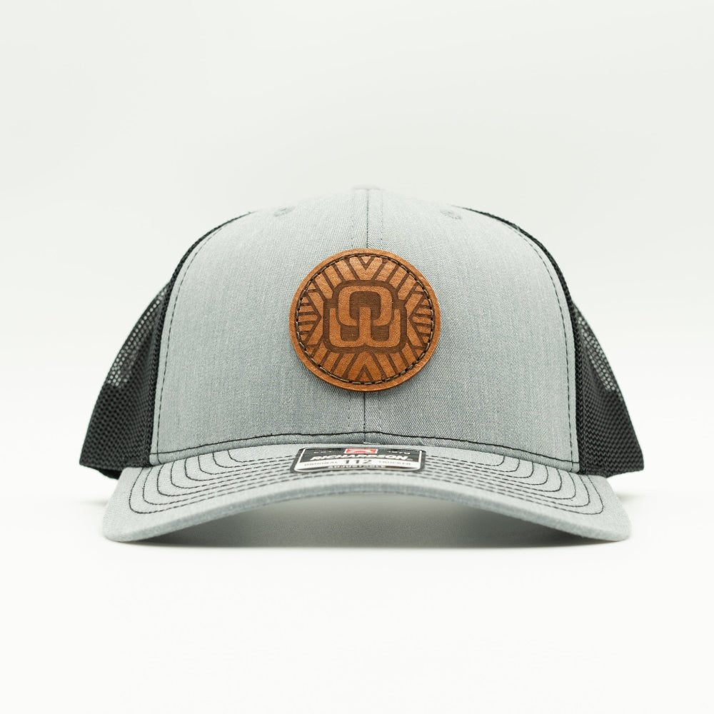 A light gray and black trucker hat with a circular brown leather patch on the front, photographed against a clean white background.

Color: Heather Grey/Black