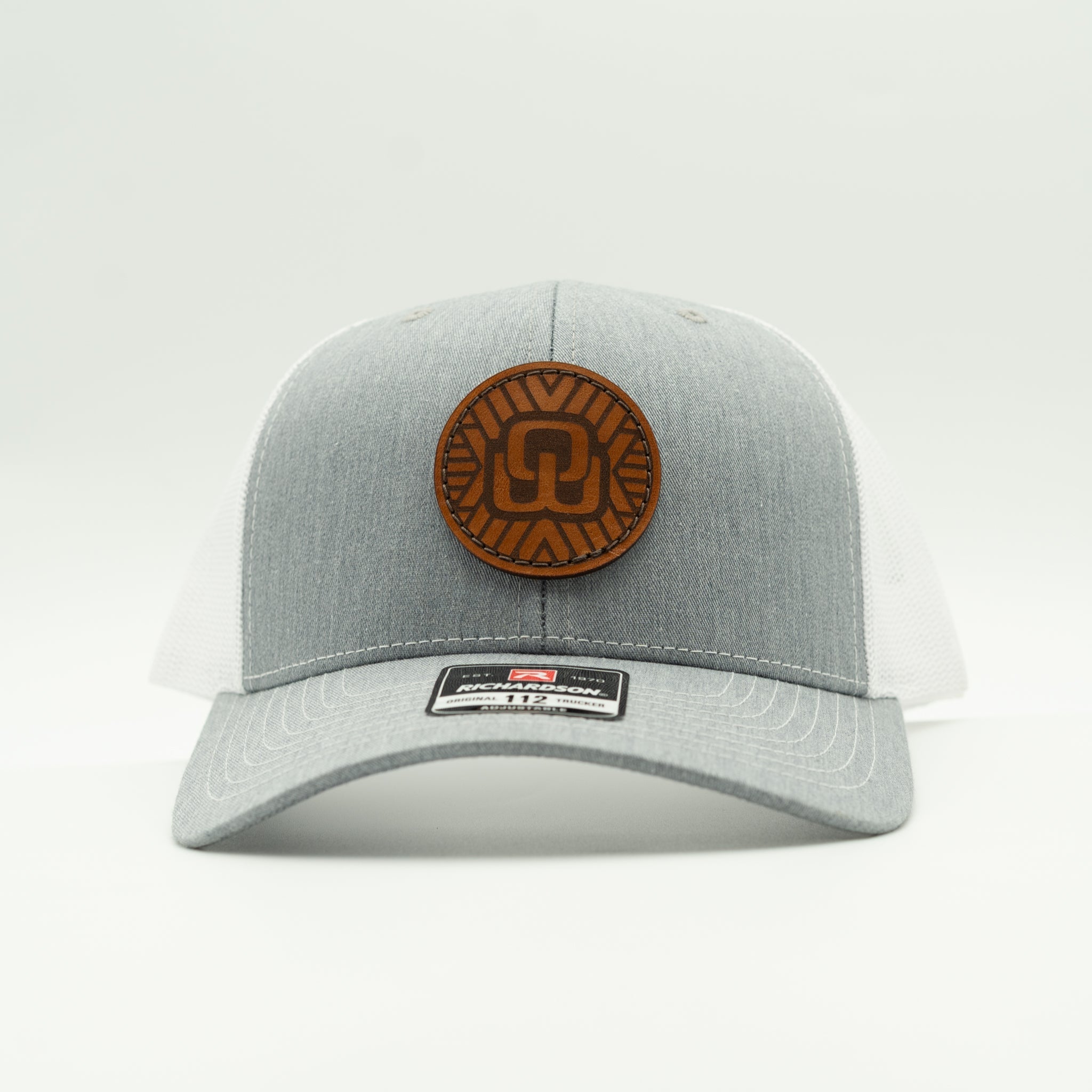 A light gray and white trucker hat with a brown circular leather patch on the front, photographed against a clean white background.

Color: Heather Grey/White