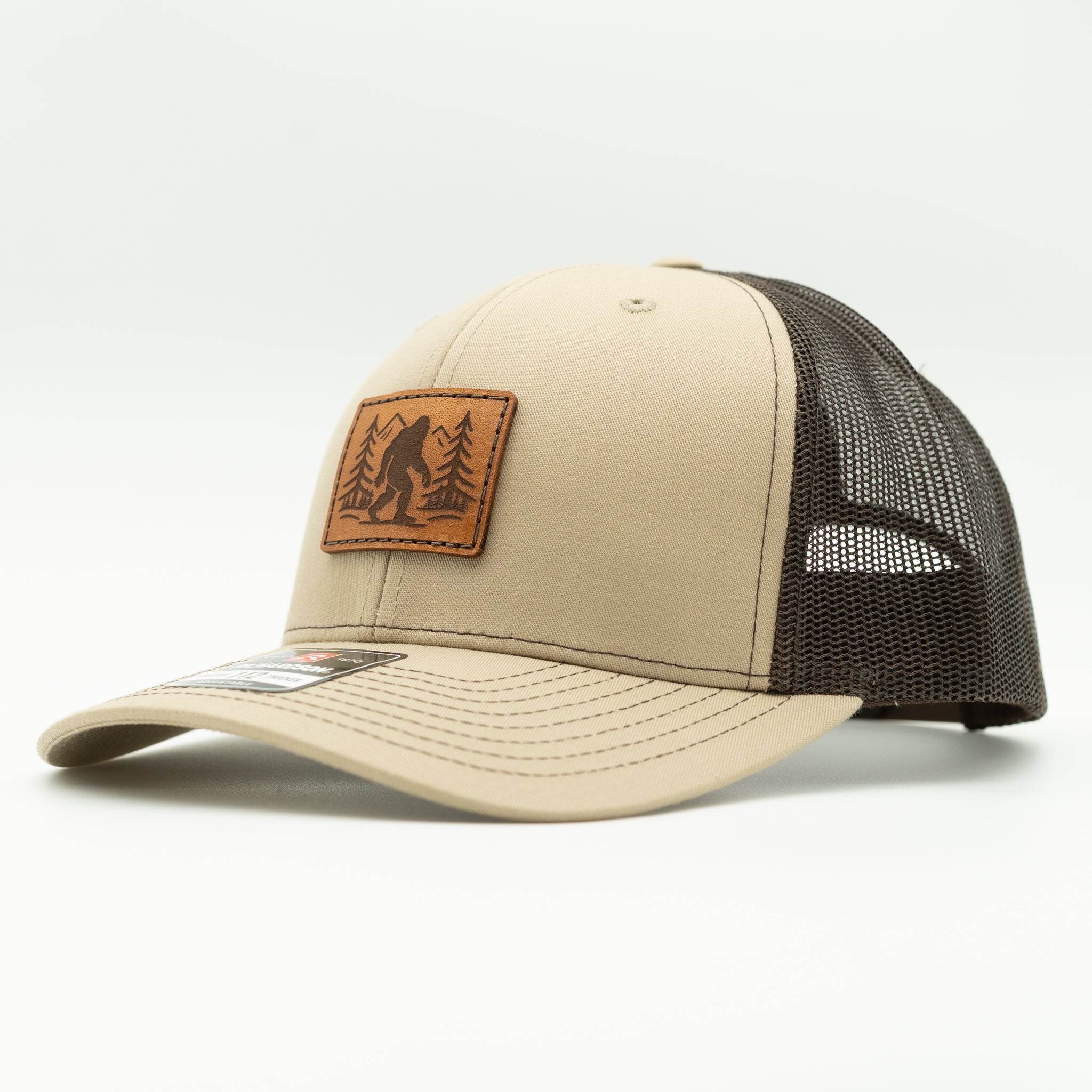 Side view of khaki front, coffee brown mesh Mountain Myth Richardson 112 hat with rectangle Bigfoot leather patch and pre-curved visor.