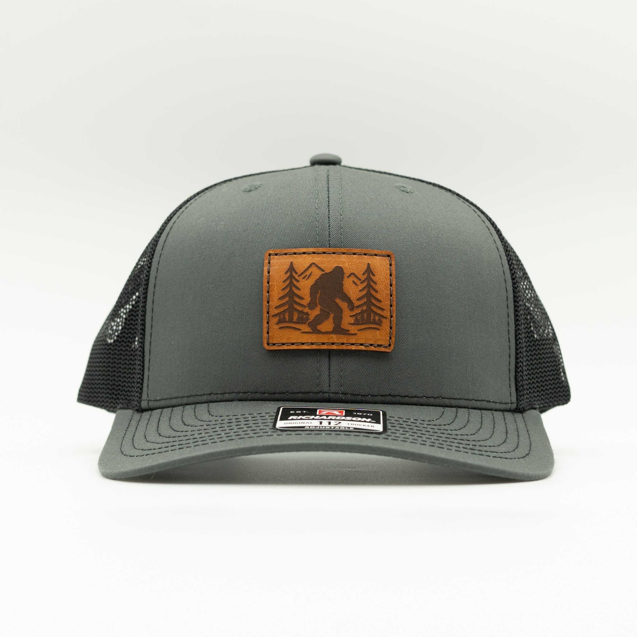 Front view of charcoal/black Mountain Myth Bigfoot leather patch trucker hat, Richardson 112 mid-profile fit with breathable mesh back.