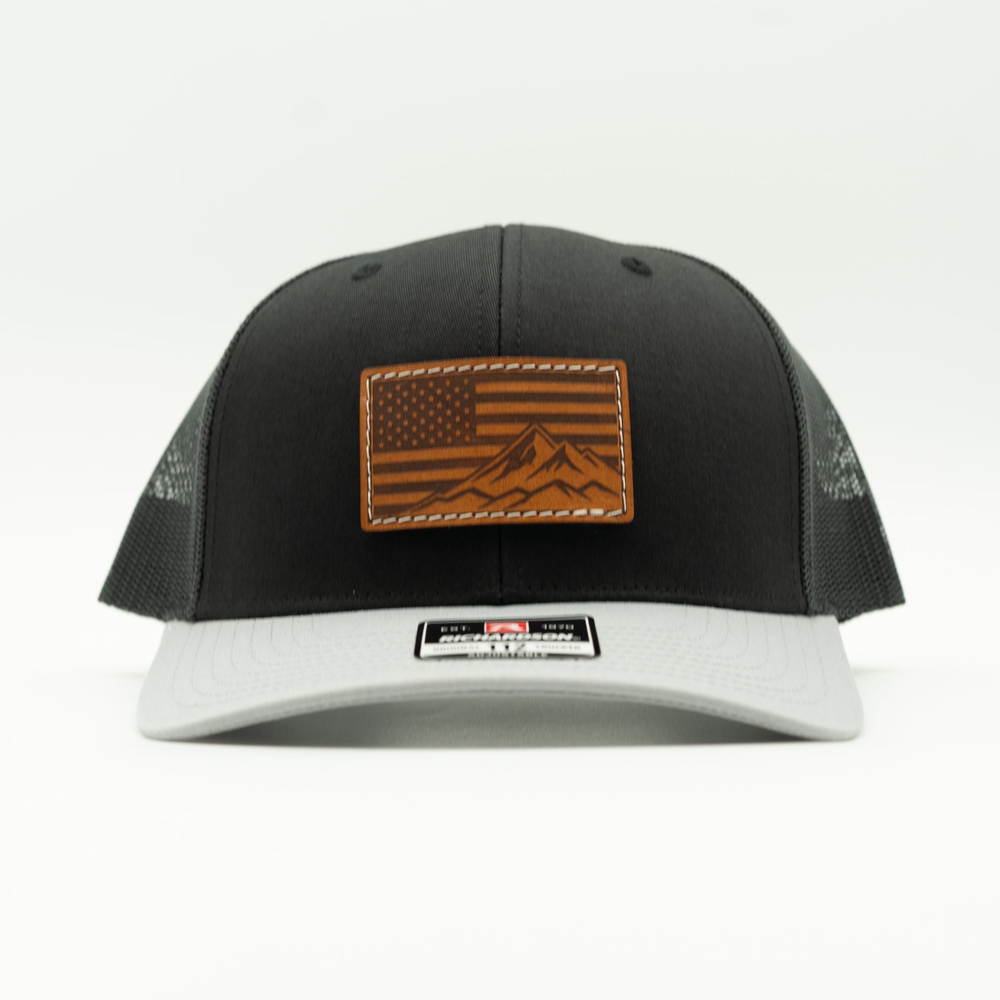 A black and gray trucker hat with a brown leather patch featuring an American flag and mountain design on the front, set against a plain white background.

Color: Black/Grey Combo
