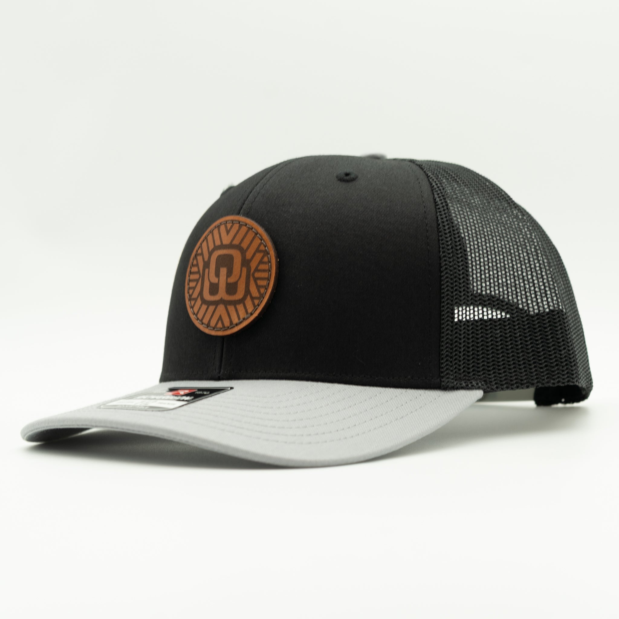 A black and gray trucker-style cap with a brown circular leather patch on the front, photographed on a clean white background.

Color: Black/Grey Combo