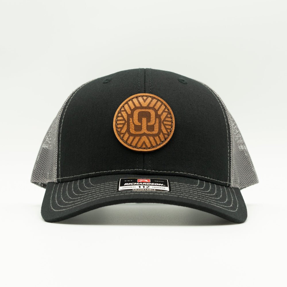 A black trucker-style baseball cap with gray mesh side panels and a circular brown leather patch on the front. A small black and white size/brand label is visible on the brim, photographed against a clean white background.

Color: Black/Charcoal