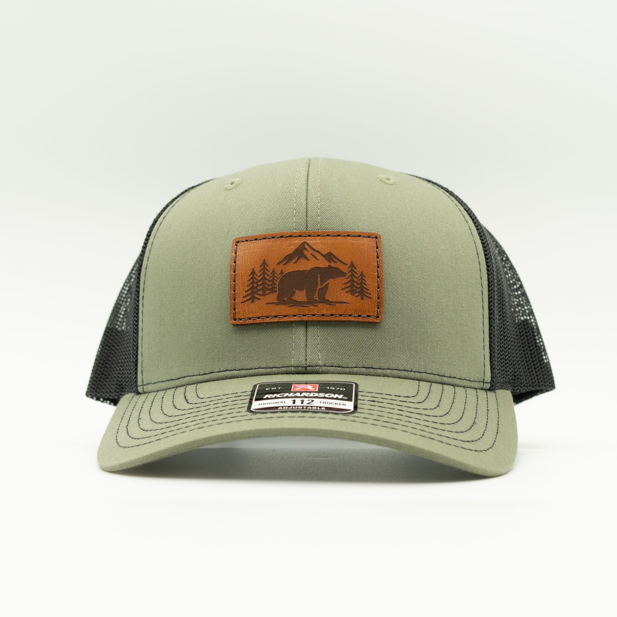 Front view of Bearline Peak leather patch hat on loden and black Richardson 112 trucker, showing bear and mountain scene patch, mid profile fit, curved bill, and mesh back.

Color: Loden/Black