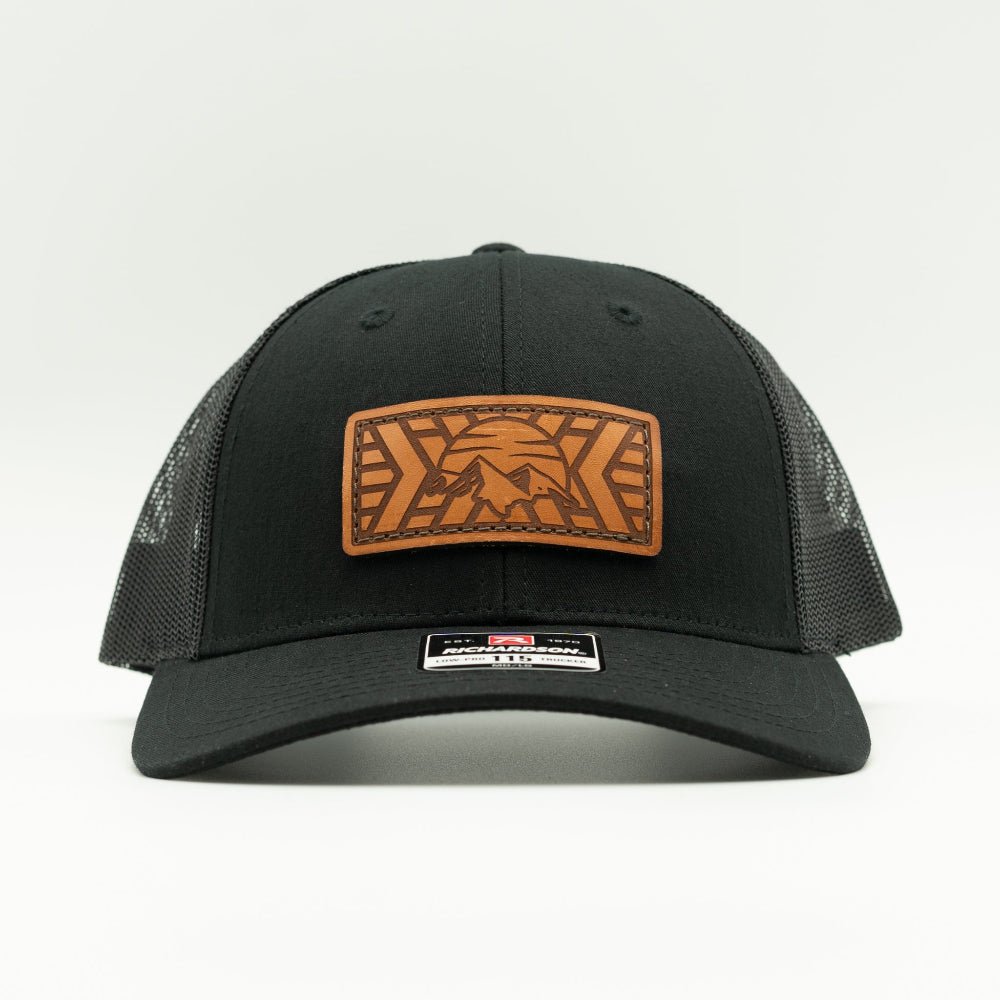 A black trucker-style cap with a brown rectangular leather patch on the front featuring a mosaic mountain design. The cap has black mesh side panels and a black curved brim, photographed against a clean white background.

Color: Solid Black