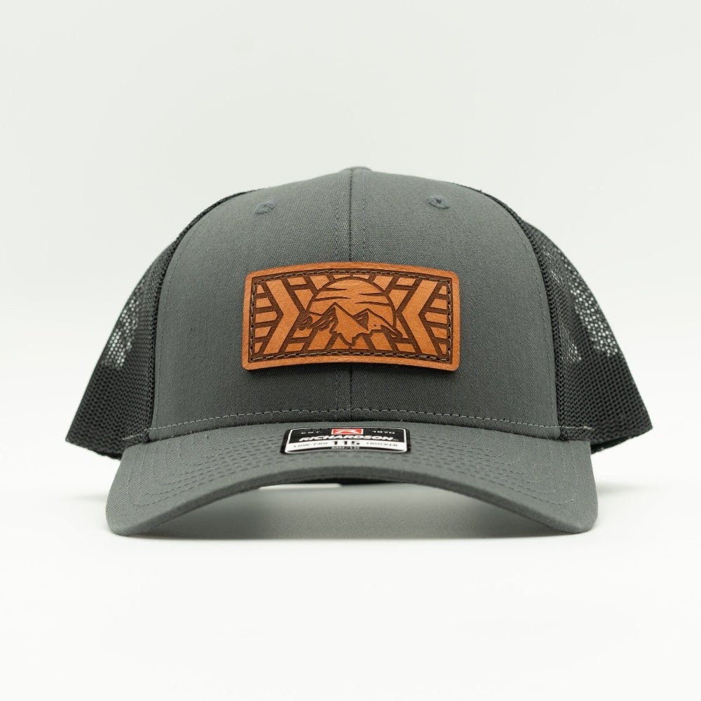A dark gray trucker hat with black mesh back panels and a brown leather patch on the front featuring a mountain and sun design.

Color: Charcoal/Black