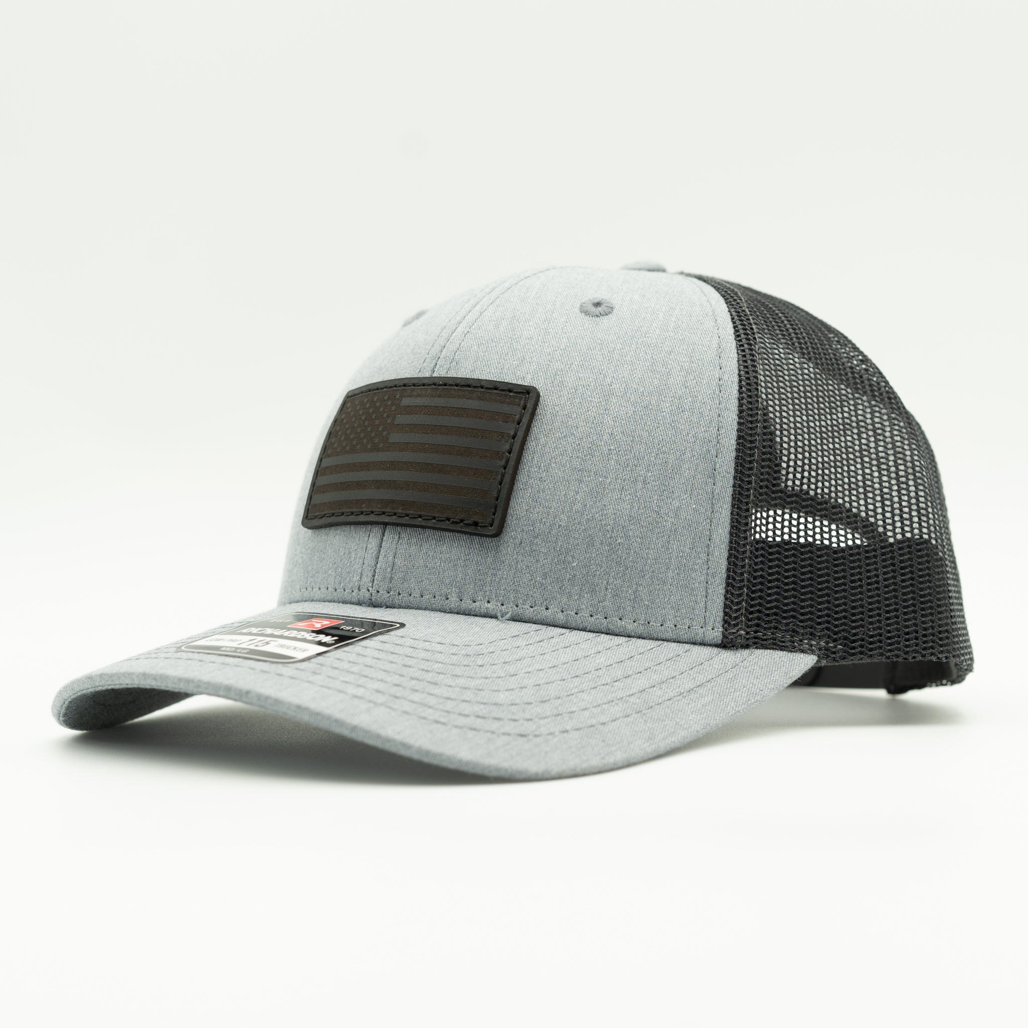 A light gray heather trucker cap with a black mesh back and a black rectangular American flag patch on the front, photographed on a clean white background.

Color: Heather Grey/Charcoal