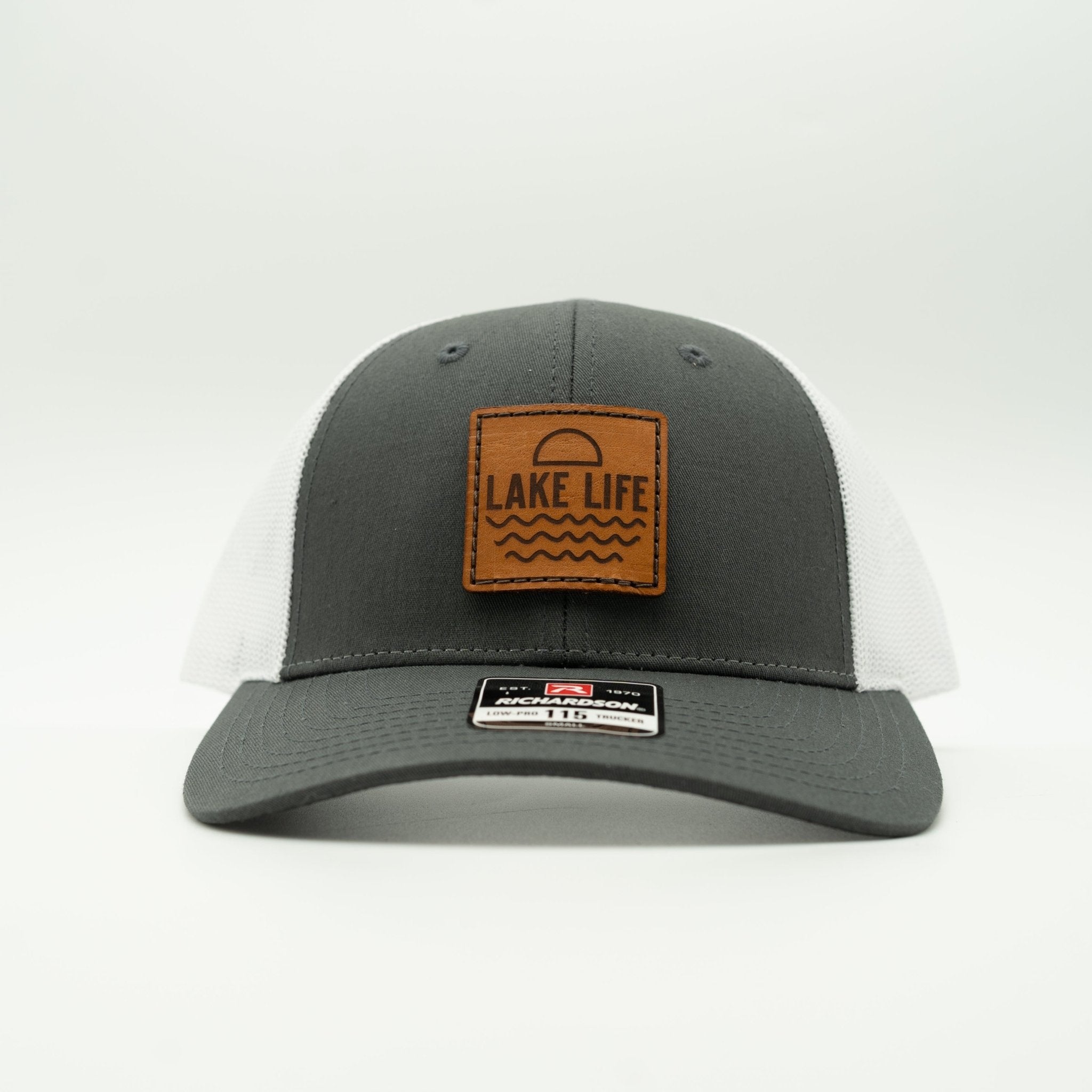 A dark gray and white trucker hat with a brown leather patch on the front that reads 'LAKE LIFE' with a sun and wave design. 

Color: Charcoal/White