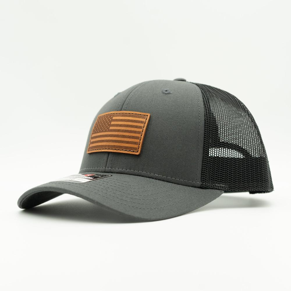A dark gray trucker-style baseball cap with black mesh back panels and a brown leather American flag patch on the front, shown on a clean white background.

Color: Charcoal/Black