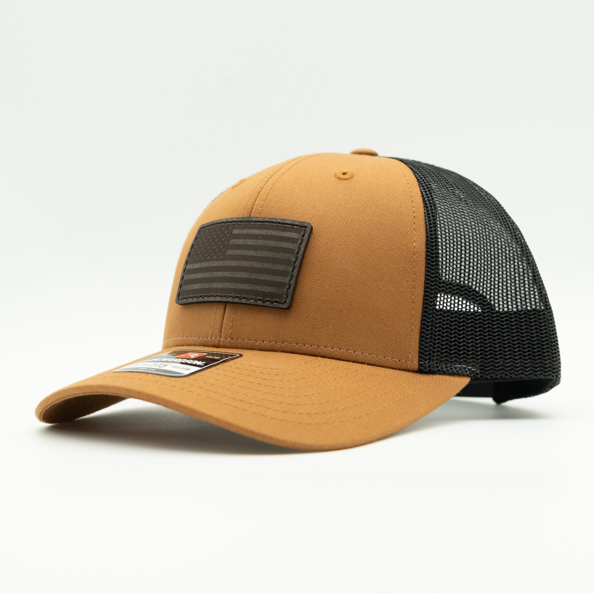 A tan and black trucker cap with a black mesh back and a black American flag patch on the front, shown on a white background.

Color: Caramel/Black