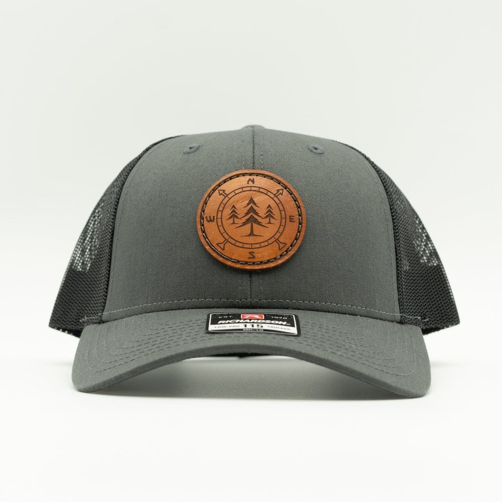 A dark gray trucker hat with black mesh side panels and a brown circular leather patch on the front featuring a tree and compass engraving.

Color: Charcoal/Black