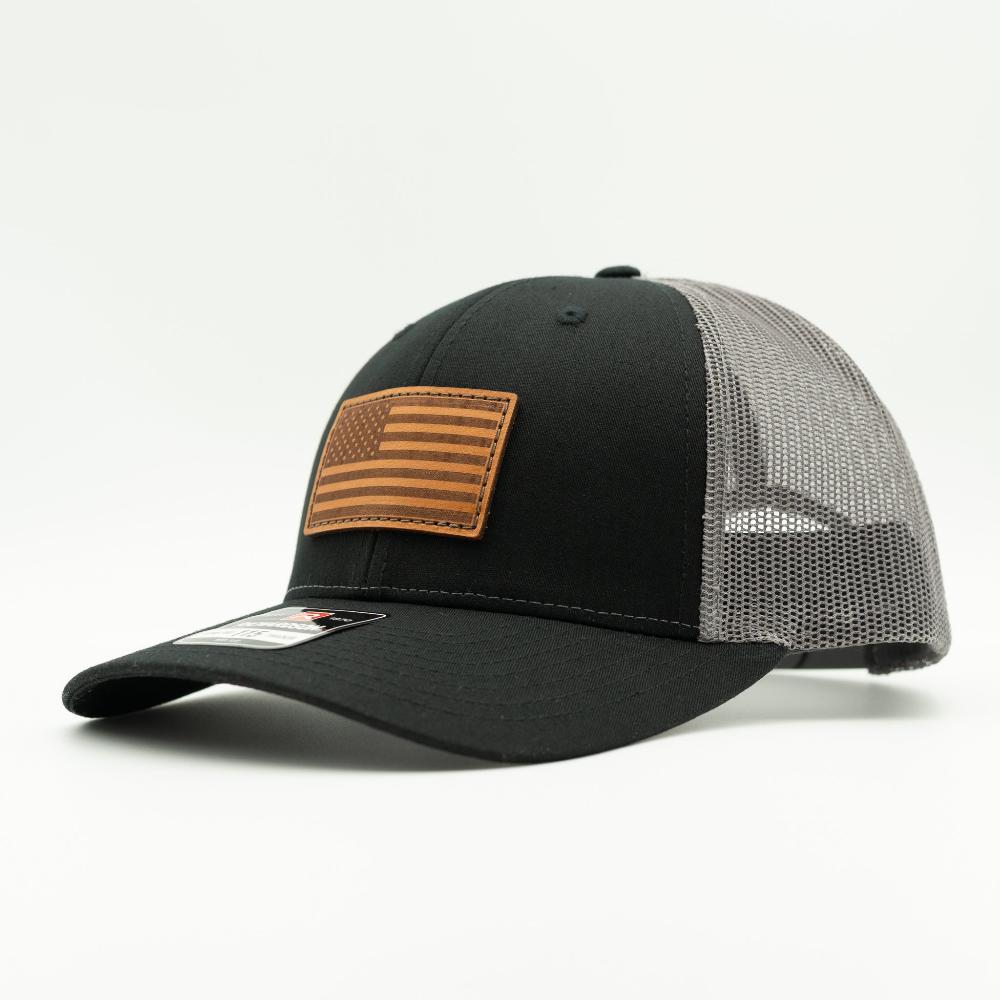 A black trucker-style baseball cap with a brown leather American flag patch on the front, featuring a black fabric front panel and a gray mesh back.

Color: Black/Charcoal