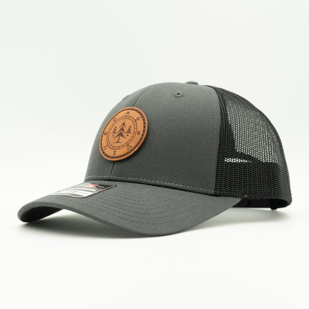 A dark gray trucker hat with black mesh back panels and a circular brown leather patch on the front featuring a pine tree and compass engraving.

Color: Charcoal/Black