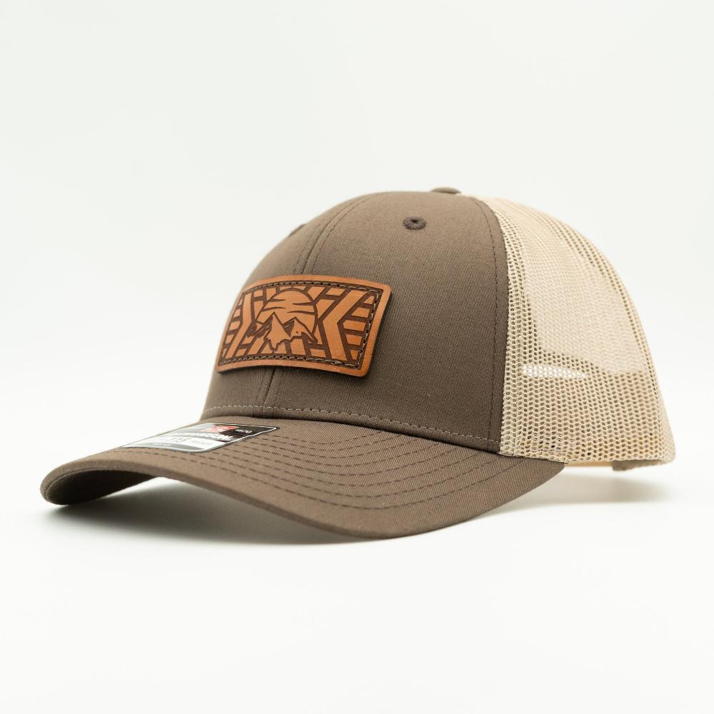 A trucker-style baseball cap with a dark brown front panel and brim, beige mesh back panels, and a brown leather patch on the front featuring a mosaic mountain patch.

Color: Brown/Khaki