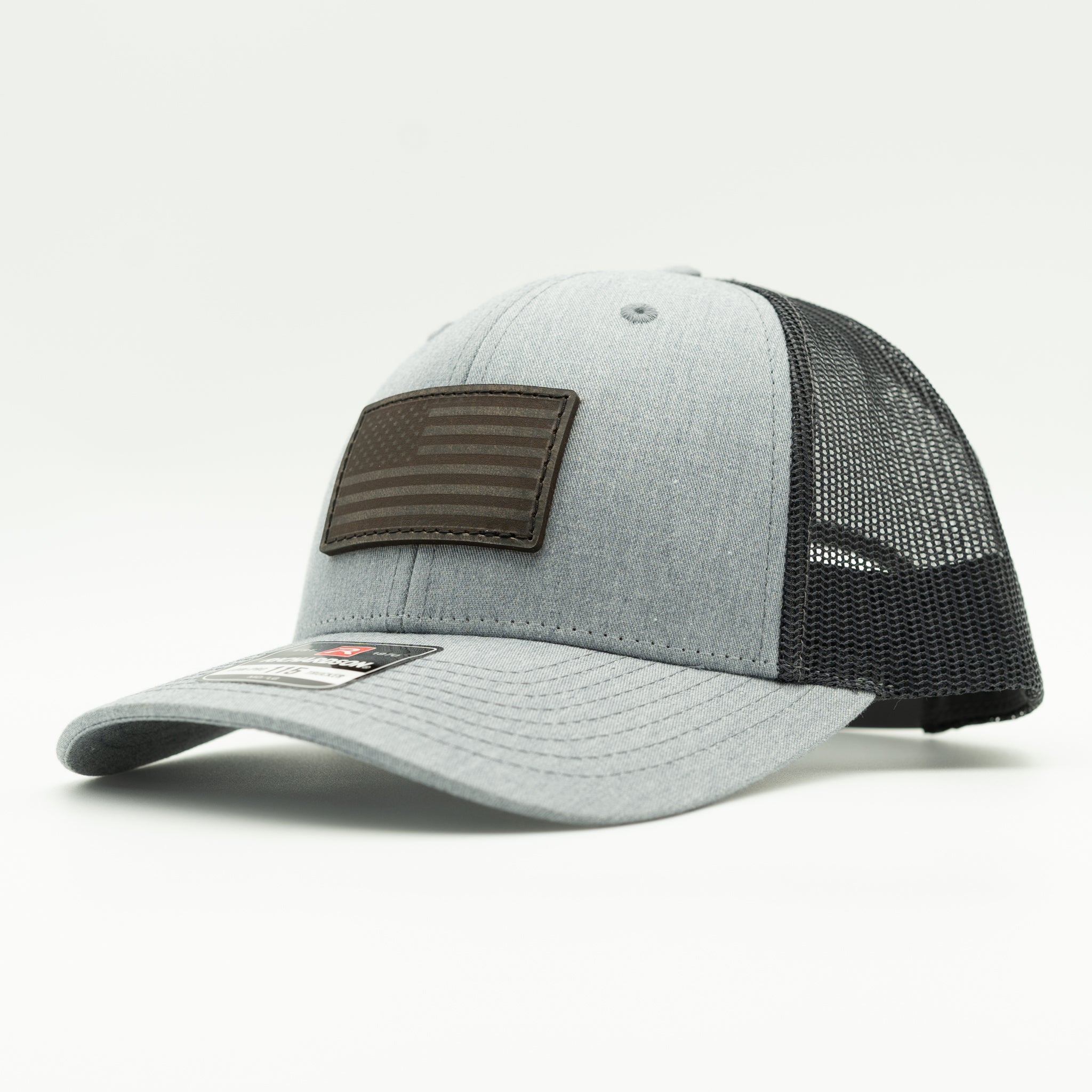 A light gray heather trucker cap with a black mesh back and a dark brown rectangular American flag patch on the front, shown on a white studio background.

Color: Heather Grey/Charcoal