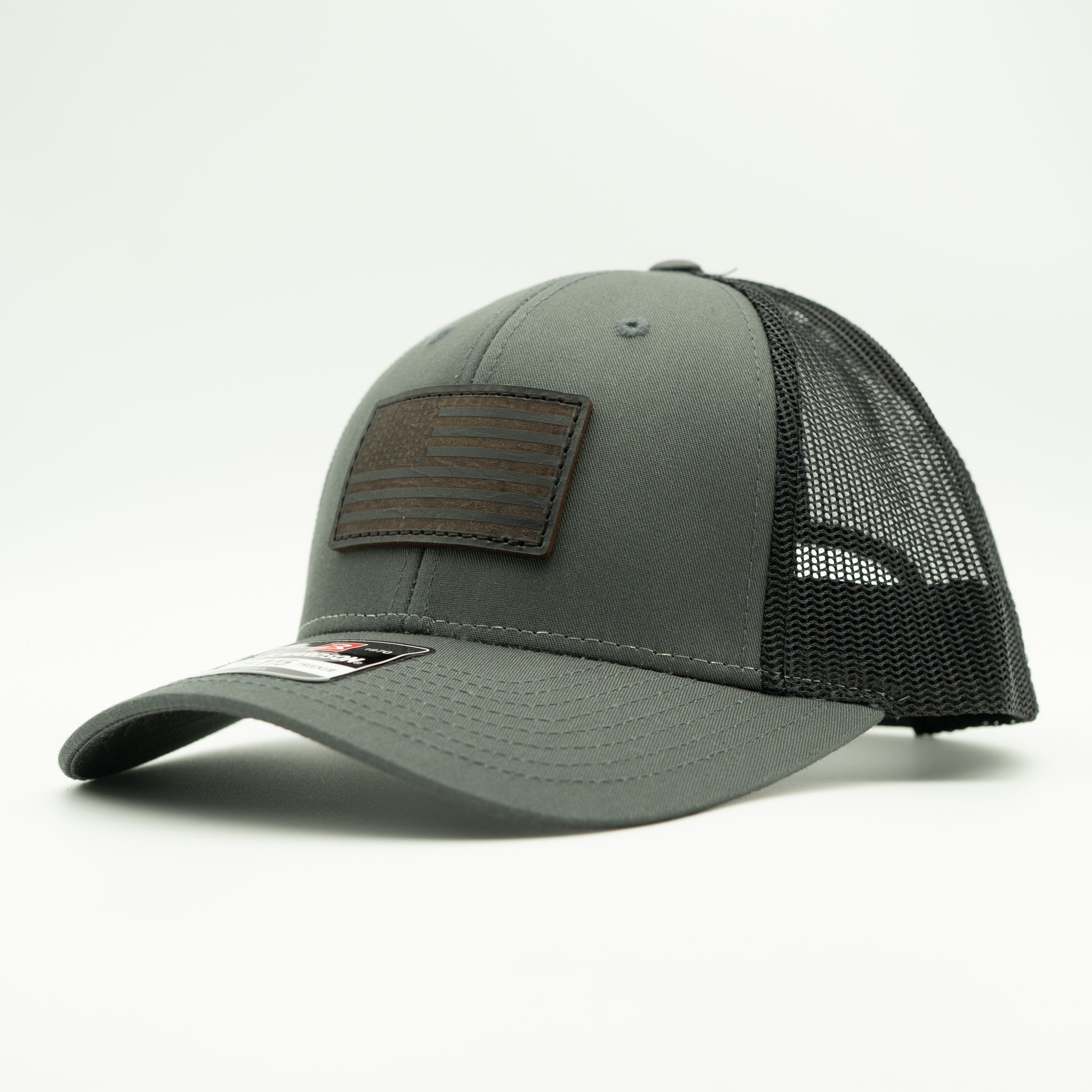 A dark gray trucker cap with a black mesh back and a brown leather American flag patch on the front, photographed on a light gray 
background.

Color: Charcoal/Black
