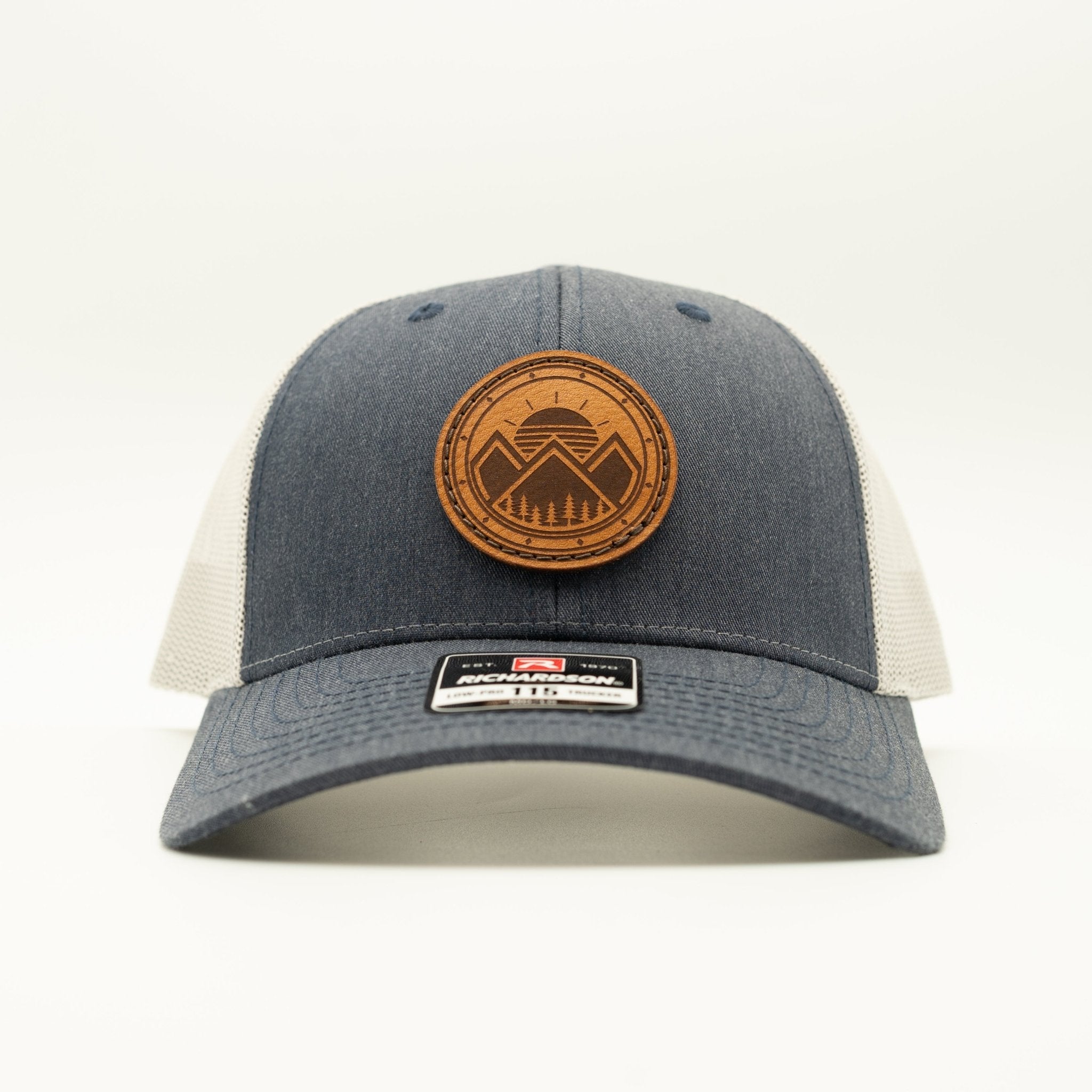 A dark gray and off-white trucker hat with a circular brown leather patch on the front displaying a mountain logo.

Color: Heather Navy/Grey