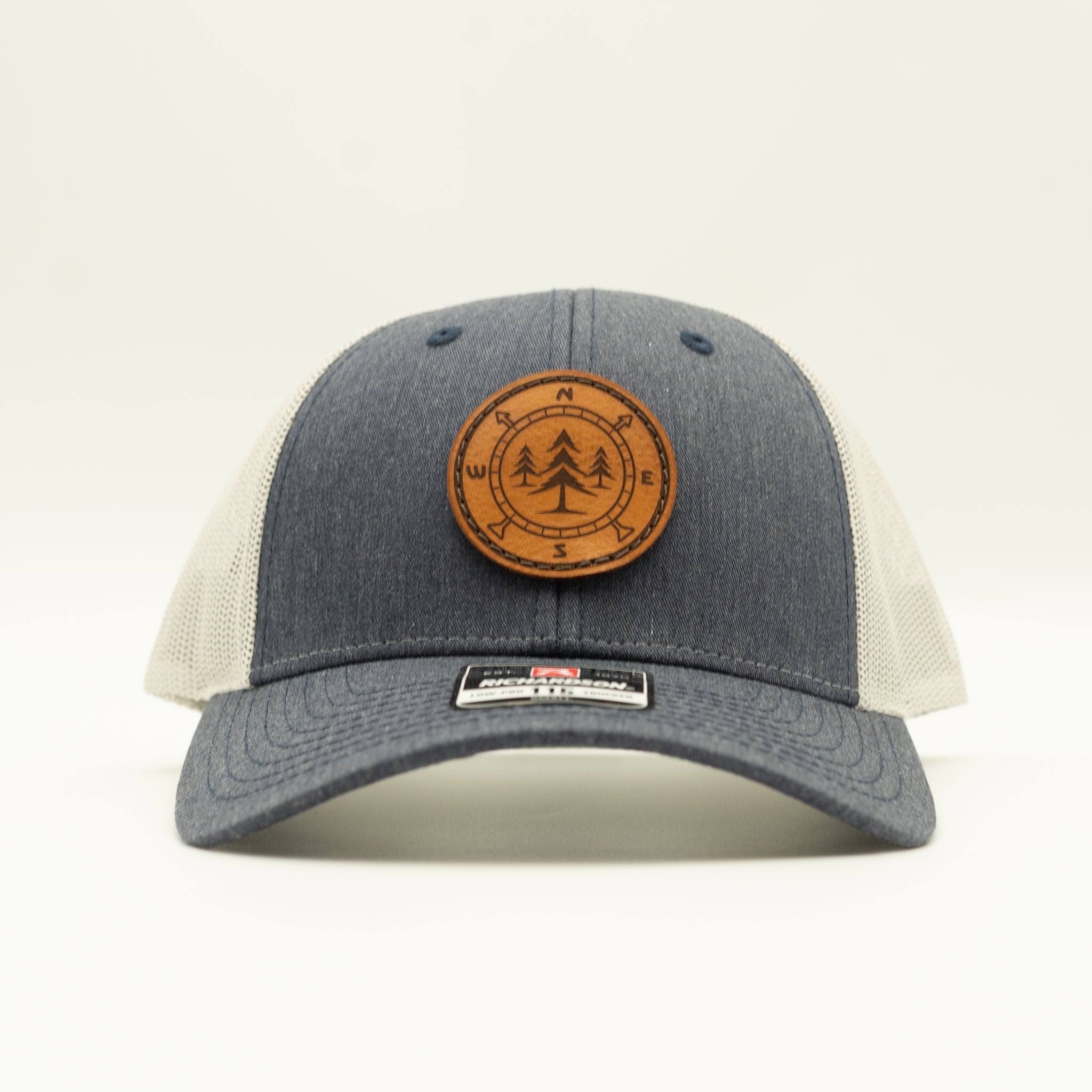 A trucker-style baseball cap with a dark gray heathered front panel and brim, off-white mesh side panels, and a circular brown leather patch on the front featuring a tree design and compass directions.

Color: Heather Navy/Grey
