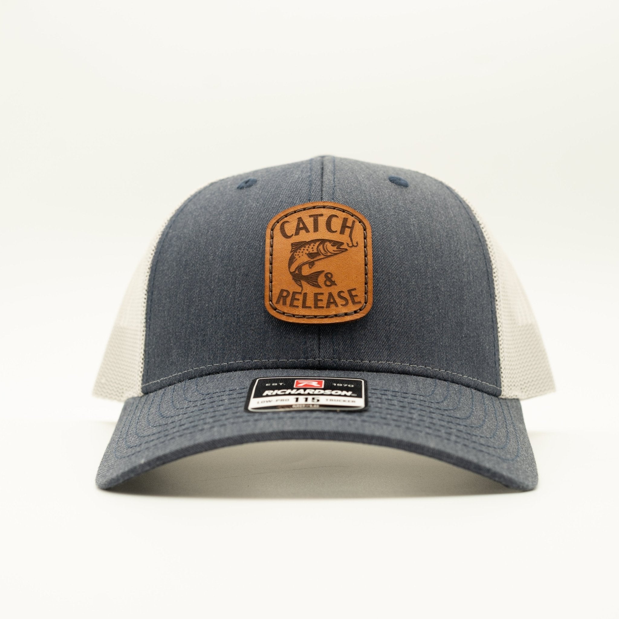 A trucker-style baseball cap with a dark gray denim front panel and brim, off-white mesh back panels, and a brown leather patch on the front that reads 'CATCH RELEASE' with a fish graphic. A small retail sticker is visible on the brim.

Color: Heather Navy/Grey