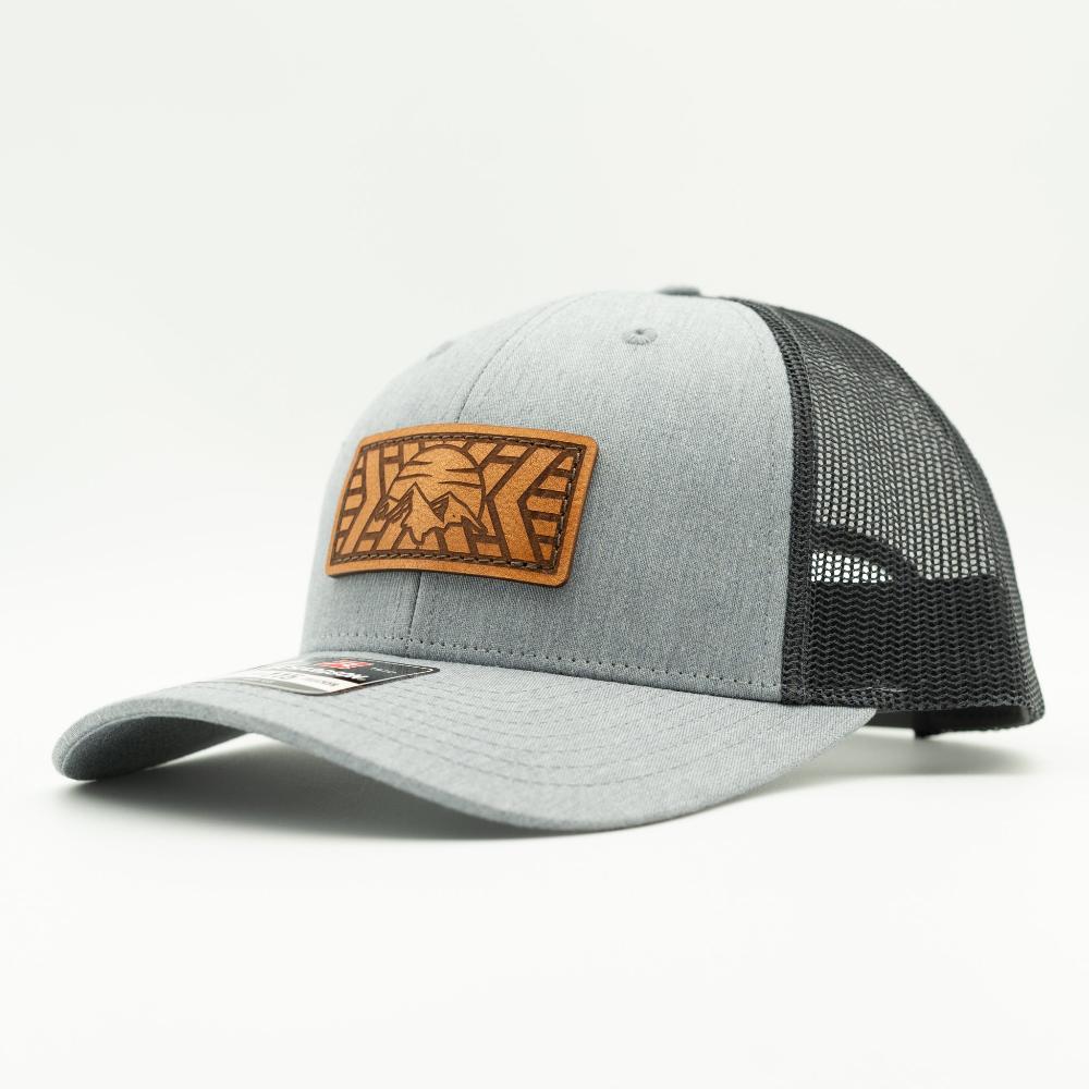 A light gray heather trucker cap with black mesh back panels and a brown leather patch on the front featuring a stylized mountain patch.

Color: Heather Grey/Charcoal